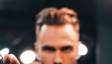 Young bearded man standing in barber shop and holding scissors.
